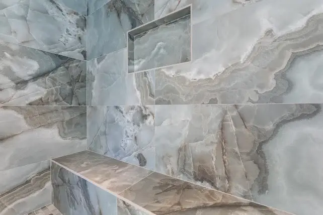 A shower with large, glossy blue-gray marble tiles featuring natural, flowing patterns. There is a built-in bench and a recessed wall niche for storage. The tiles create a luxurious and modern appearance.