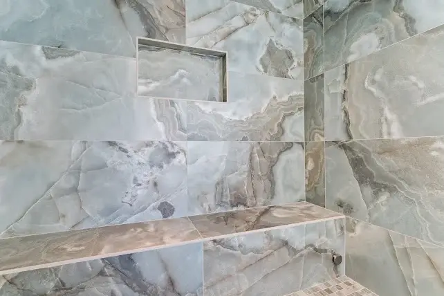 A shower with walls and a built-in bench covered in large, glossy, marble-patterned tiles in shades of gray and blue; a recessed niche for storage is set into the wall above the bench.