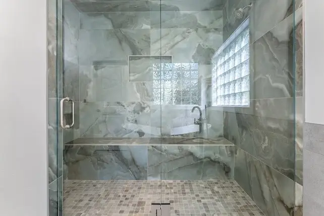 Spacious modern shower with glass door, gray and white marble-patterned tiles, built-in bench, a small window of glass blocks, and a rainfall showerhead.