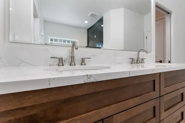 Close-up of a modern bathroom vanity with a white marble countertop, dual sinks, stainless steel faucets, and brown wooden cabinets, with a large mirror above and a glimpse of a shower and door in the background.