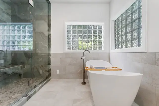 Modern bathroom with a freestanding white bathtub, a wooden bath tray, glass-enclosed shower, light gray tile walls and floor, and two large glass block windows letting in natural light.