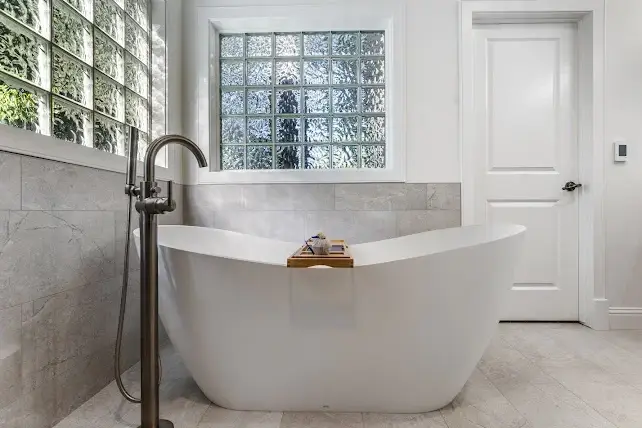 Modern bathroom with a sleek white freestanding bathtub, a floor-mounted faucet, light gray tiled walls and floor, glass block windows, and a white door. A wooden bath tray rests across the tub.