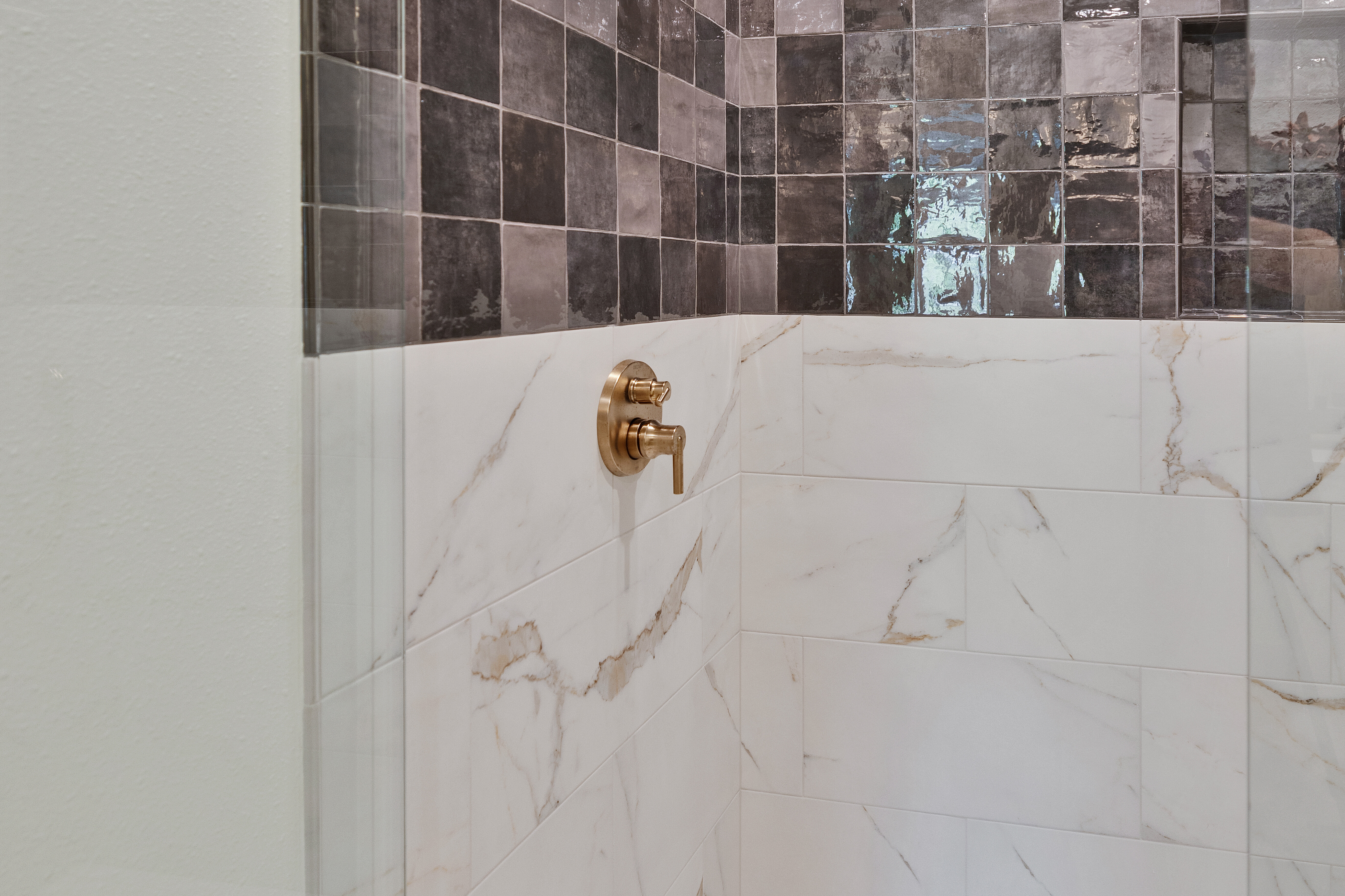 Modern shower with gold hardware, white marble lower tiles, and glossy dark gray upper tiles. Glass door is partially visible on the left side. Floor is white with a square drain.