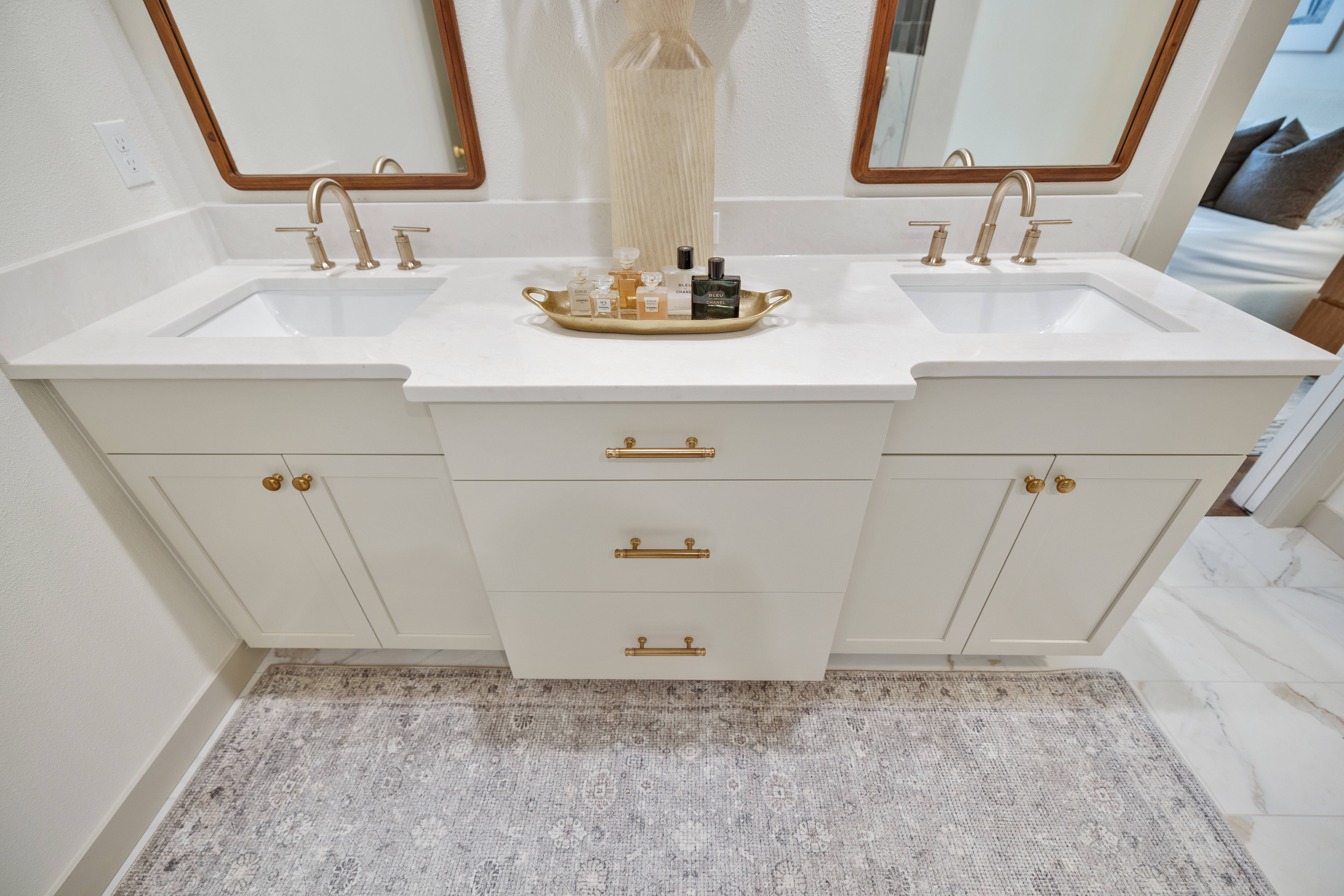 White double bathroom vanity with gold hardware, two sinks, and two mirrors. A decorative tray with toiletries is placed in the center. A neutral rug and marble-like flooring are visible below.