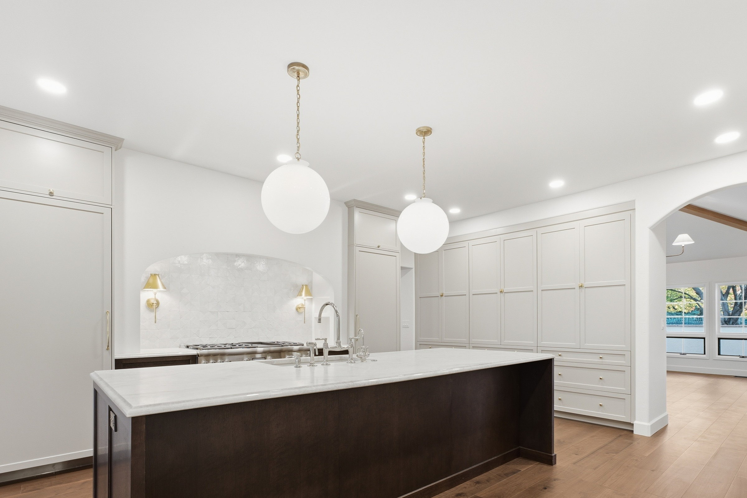 Remodeled kitchen with a dark stained island, marble countertops, globe pendants, an arched range alcove and white oak floors.