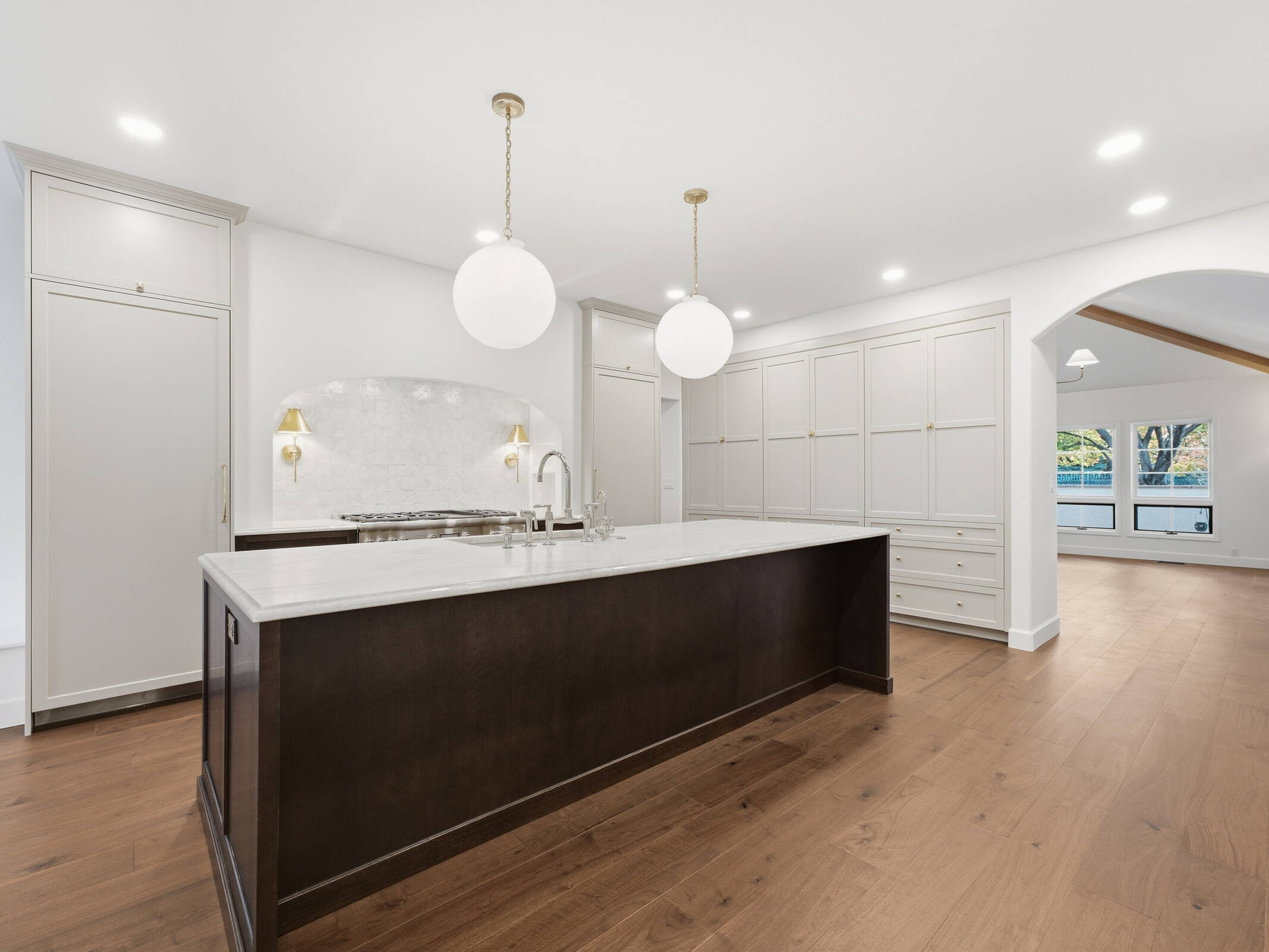 Remodeled kitchen with a dark stained island, white marble countertop, globe pendants and an arched stone range alcove with brass sconces.