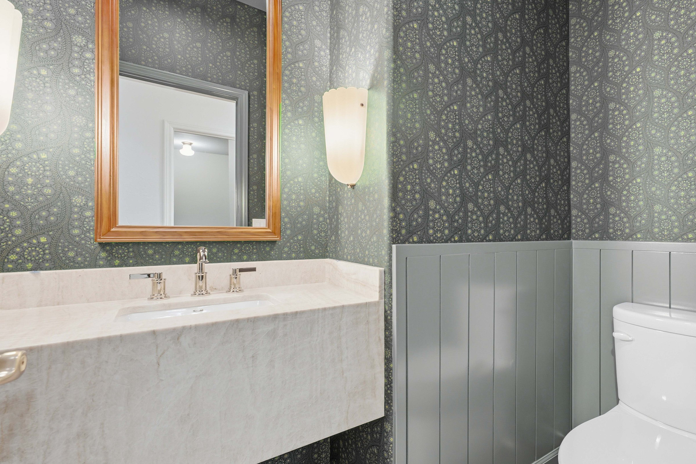 Remodeled powder bathroom with dark botanical wallpaper, painted wainscot, a marble vanity and a gold framed mirror.