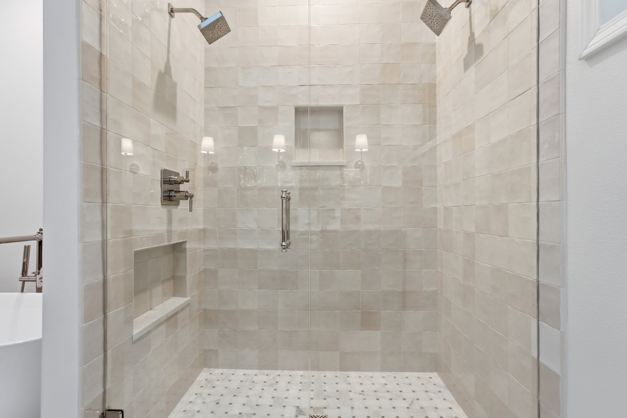 Remodeled walk in shower lined in handmade look tile with two shower heads, a recessed niche and a marble mosaic floor.